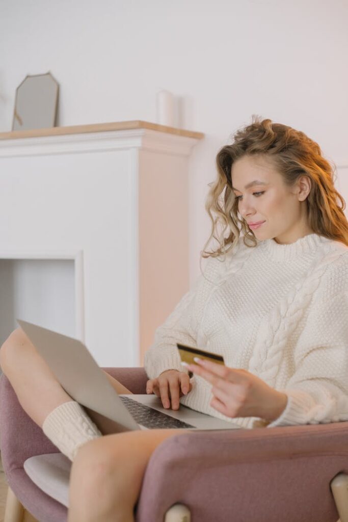 Adult woman in cozy sweater shopping online using laptop and credit card during daytime.